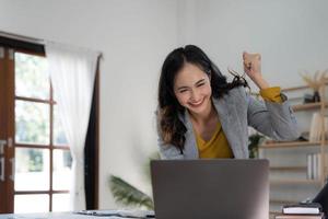 une femme asiatique heureuse et excitée célèbre avec un ordinateur portable, un succès ou une action heureuse. indépendant ou entrepreneur utilisant la technologie et démarrant une maison moderne. photo