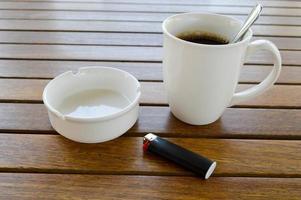 une tasse en céramique blanche avec un café chaud rafraîchissant le matin avec une boisson au thé et une cuillère à thé brillante et un cendrier vide avec un allume-cigare noir se dresse sur une table en bois dans un café photo