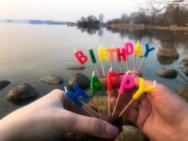 inscription de joyeux anniversaire faite de bougies de vacances entre les mains d'un homme et d'une femme en face de l'eau de la rivière du lac océanique photo
