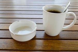 une tasse en céramique blanche avec un café chaud rafraîchissant le matin avec une boisson au thé et une cuillère à café brillante de thé et un cendrier de cigarette vide est sur une table en bois dans un café photo