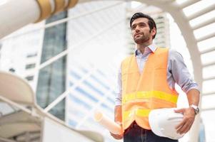 jeune homme caucasien tenant un gros papier, un gars portant une chemise bleu clair et un jean avec un gilet orange et un casque blanc pour la sécurité dans la zone de construction. photo