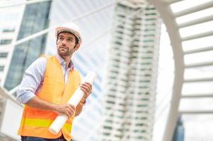 jeune homme caucasien tenant un gros papier, un gars portant une chemise bleu clair et un jean avec un gilet orange et un casque blanc pour la sécurité dans la zone de construction. photo