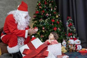 bébé caucasien est présent du père noël la nuit par l'arbre de noël entièrement décoré pour le concept de célébration de la saison photo