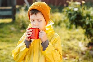 un petit garçon en imperméable jaune boit dans une tasse rouge dans la rue. notion de camping. photo d'automne avec une tasse de cacao dans la rue