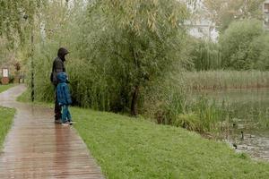 un homme avec un enfant dans une veste à capuche, se tenir debout et regarder le lac un jour de pluie photo