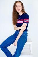 portrait d'une jeune fille souriante assise sur une chaise, sur fond blanc, aux cheveux longs, dans un t-shirt moulant et un jean photo