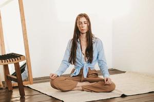 fille aux cheveux longs et noirs assis pose de lotus méditant sur l'intérieur de la salle lumineuse au sol. pratique du yoga photo