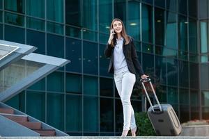 jolie hôtesse de l'air souriante transportant des bagages allant à l'avion à l'aéroport. femme d'affaires à l'aéroport international regardant son téléphone portable photo