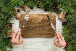 vue de dessus la fille tient une fourchette et un couteau à la main et est prête à manger. plaque rectangulaire en bois vide sur fond de noël en bois. concept de plat de dîner de vacances avec un décor de nouvel an photo