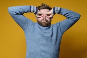 un homme bizarre, barbu et hirsute, vêtu d'un chandail, pressait les doigts contre le visage comme s'il s'agissait de lunettes. fond jaune. photo