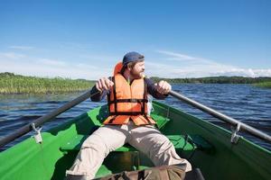 homme barbu satisfait dans un gilet de sauvetage ramant avec des rames sur un bateau et profitant du magnifique paysage. concept de mode de vie actif. photo