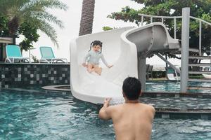 père et fille asiatiques nageant en jouant au toboggan dans la piscine du complexe, souriant et riant. s'amuser dans la piscine de l'hôtel de villégiature, concept familial heureux. photo