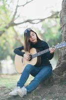 des musiciennes asiatiques portant une chemise noire à manches longues et des lunettes de soleil sur la tête. chanter et jouer de la guitare acoustique sous un arbre dans le parc le matin. photo