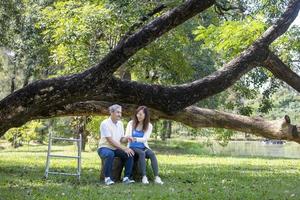 père senior asiatique avec marcheur et fille assis ensemble sous le grand arbre dans le parc pendant l'été pour un exercice léger et un concept de thérapie physique photo