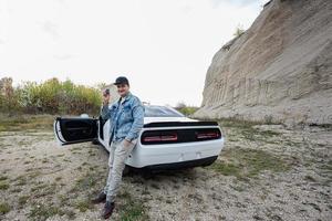 heureux nouveau propriétaire homme en veste de jeans et casquette se tient près de son rêve - muscle car blanc. montrer les clés. photo