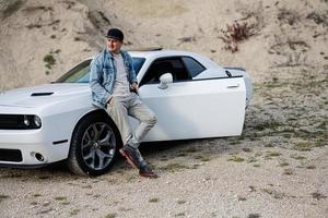 bel homme en veste de jeans et casquette se tient près de sa voiture de muscle blanche en carrière. photo