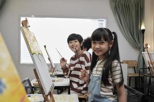 portrait d'enfants asiatiques regardant la caméra et souriant avec une image en couleur acrylique peinte sur toile dans une salle de classe d'art et un apprentissage créatif avec talents et compétences dans l'enseignement en studio de l'école primaire. photo