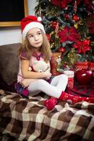 une petite fille tenant un ours en peluche, assise sur une couverture à carreaux dans les décorations de noël près d'un sapin de noël avec des boîtes de cadeaux et un bonnet de noel. nouvel an, jeu d'enfants photo