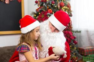 petite fille en bonnet de noel sur les genoux du père noël près du sapin de noël dans un décor de noël. montre une boîte avec un cadeau, une peluche, touche sa barbe et rit. nouvel an, grand-père russe givre photo