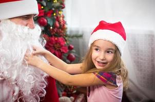 petite fille en bonnet de noel sur les genoux du père noël près du sapin de noël dans un décor de noël. montre une boîte avec un cadeau, une peluche, touche sa barbe et rit. nouvel an, grand-père russe givre photo