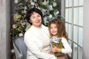 grand-mère avec une petite fille sur fond de décorations de noël et une grande fenêtre. vacances en famille, émotions, coffret cadeau. petite-fille sur les genoux de grand-mère. nouvel An photo