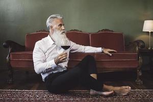 bel homme âgé barbu assis sur le sol et buvant du vin rouge photo