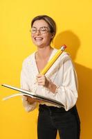 femme gaie portant des lunettes tenant un gros crayon et un cahier sur fond jaune photo