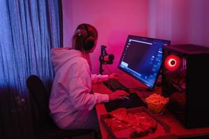 femme gamer ou développeur de logiciels assis devant l'ordinateur personnel moderne et mangeant de la malbouffe la nuit dans la chambre avec des néons photo