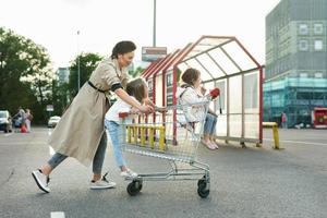 heureuse mère et ses filles s'amusent avec un panier sur un parking à côté d'un supermarché. photo