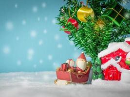 joyeux père noël avec une boîte de cadeaux sur le traîneau à neige allant à la maison. près de la maison ont bonhomme de neige et arbre de Noël. père noël et maison sur la neige. concept de noël et bonne année. photo