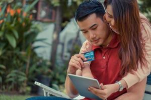 heureux jeune couple assis dans le jardin et tenant une tablette et une carte de crédit factice et achetant actuellement des produits via le commerce électronique. concept de shopping sur internet avec paiement par carte de crédit. photo