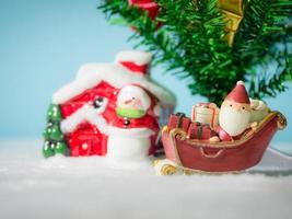 joyeux père noël avec une boîte de cadeaux sur le traîneau à neige allant à la maison. près de la maison ont bonhomme de neige et arbre de Noël. père noël et maison sur la neige. concept de noël et bonne année.