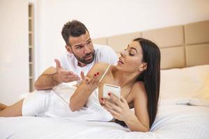 photo d'un jeune couple au lit, une femme utilise un smartphone, essaie d'expliquer l'envie d'un mari en colère, elle n'a pas un autre homme pointant l'écran de la main, se sent confus. mari jaloux espionnant le téléphone portable de sa femme