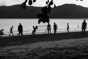 image monochrome de silhouette groupe de personnes jouant au football sur la plage au coucher du soleil avec paysage marin et montagne en arrière-plan. photo