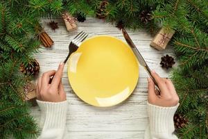 vue de dessus la fille tient une fourchette et un couteau à la main et est prête à manger. assiette vide en céramique ronde sur fond de noël en bois. concept de plat de dîner de vacances avec un décor de nouvel an photo