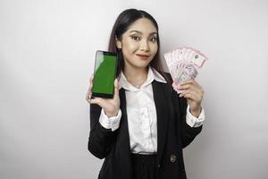 une jeune femme d'affaires heureuse porte un costume noir, montrant son téléphone et son argent en roupie indonésienne isolée par fond blanc photo