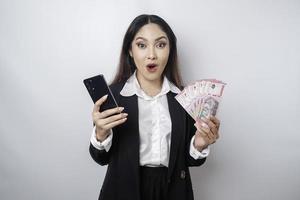 femme d'affaires asiatique surprise portant un costume noir tenant son smartphone et de l'argent en roupie indonésienne, isolée sur fond blanc photo