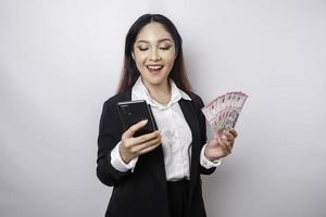une jeune femme d'affaires heureuse porte un costume noir, tenant son téléphone et de l'argent en roupie indonésienne isolée par fond blanc photo