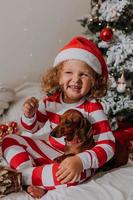 chien et petite fille en pyjama rouge et blanc mange une savoureuse sucette de noël maison assis dans son lit. enfant et un animal de compagnie. bébé et teckel s'amusent et célèbrent la nouvelle année. mode de vie photo