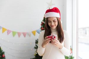 femme brune souriante en pull et bonnet de noel tenant une petite boîte cadeau et se réjouit de la décoration de noël fond blanc photo