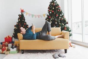 un homme seul semble seul en utilisant son téléphone et s'allonge sur un canapé dans le salon décoré d'arbres de noël et de coffrets cadeaux photo