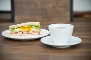 sandwich et tasse de café sur une assiette blanche et une table en bois, le sandwich est fait de pain, de saucisses, d'œufs au plat et de légumes est un petit-déjeuner américain ou la restauration rapide est une nourriture malsaine ou de la malbouffe photo