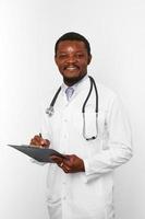 homme médecin barbu noir souriant en robe blanche avec stéthoscope remplissant les dossiers médicaux sur le presse-papiers photo