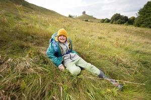 garçon souriant assis sur la colline d'automne portant une veste verte, un chapeau jaune, un sac à dos et des bottes en caoutchouc. photo