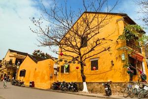 hoi an, vietnam - 12 février 2018. rue touristique de la vieille ville avec maisons jaunes, grand arbre, motos et vélos. photo