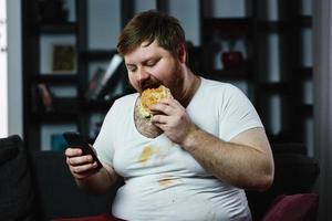 gros homme vérifie quelque chose dans son smartphone pendant qu'il mange un hamburger photo