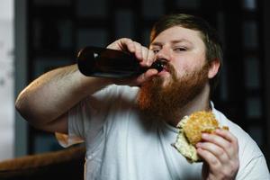 gros homme regarde la télévision, mange un hamburger et boit de la bière photo