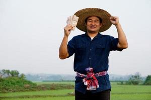 un agriculteur asiatique heureux est à la rizière, détient de l'argent en billets de banque thaïlandais. concept, un agriculteur heureux obtient des bénéfices, des revenus, de l'agriculture soutenant l'argent. fier sur les récoltes. photo