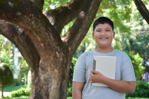 portrait jeune garçon asiatique tient laplet et se tient sous le grand arbre, mise au point douce, concept d'utilisation de la technologie dans la vie quotidienne. photo