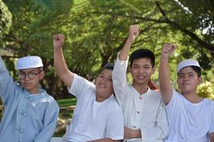 les jeunes garçons musulmans asiatiques lèvent la main droite pour montrer joyeusement leur victoire et leur succès sous un arbre dans le parc de l'école, mise au point douce et sélective. photo